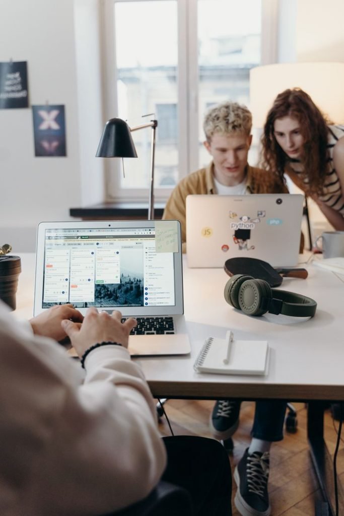 man-and-woman-working-together-7439137 Young team collaborating at a modern workspace with laptops and headphones.